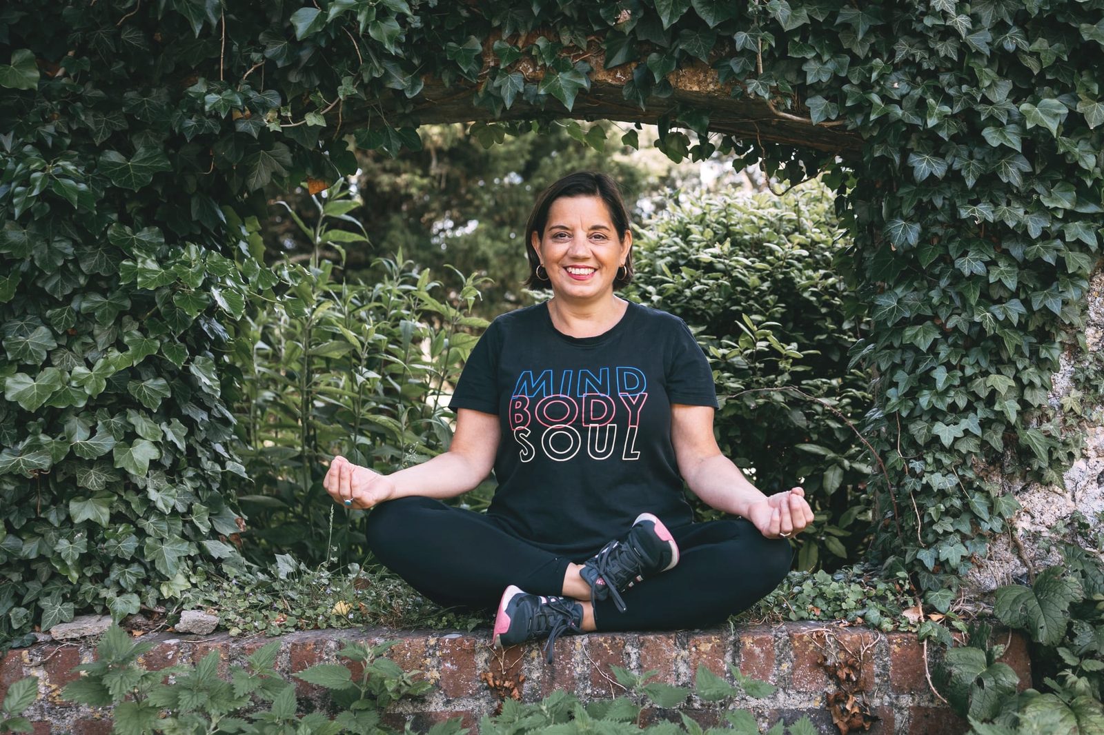 Celine sitting cross-legged in a leafy ivy doorway on an old brick wall, smiling, wearing a black Mind Body Soul t-shirt