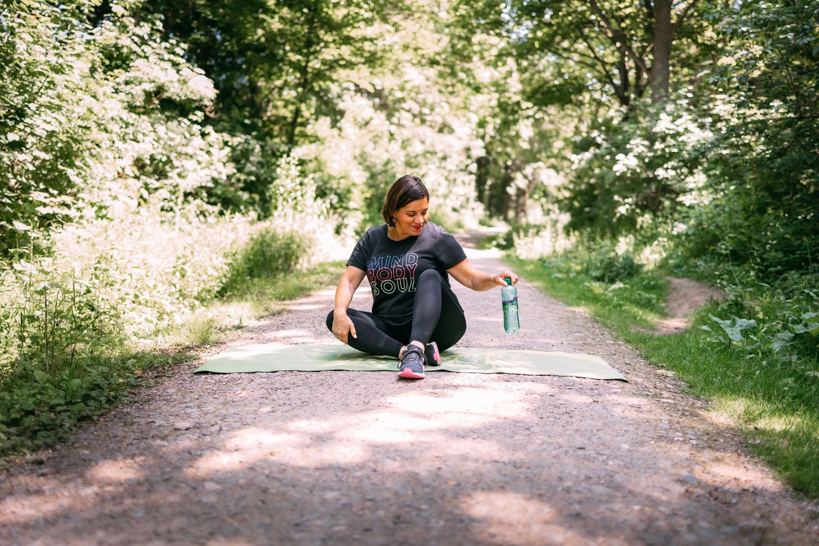 Celine sitting on a green mat on a sunlit woodland path, holding a water bottle