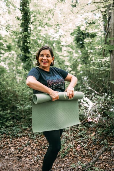 Celine laughing in a sunlit wood, holding a rolled green exercise mat